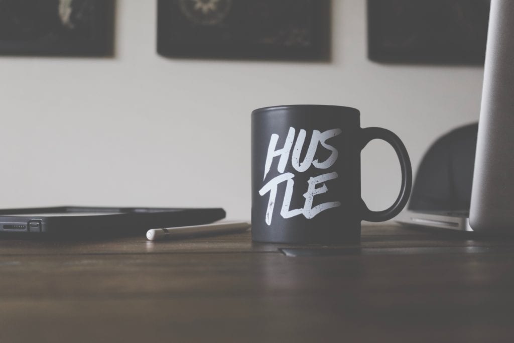 Black coffee mug sitting on a desk with the word "hustle" on it. The call to "hustle" is an example of the pursuit of a faulty view of success.